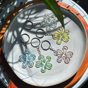 May include: Five flower-shaped keychains made of translucent, iridescent material. Each keychain features a silver-toned metal split ring attached by a short chain. The flower charms display shifting colors including clear, yellow, blue, green, and pink.