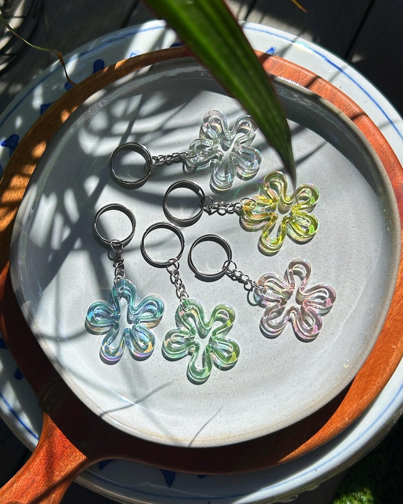 May include: Five flower-shaped keychains made of translucent, iridescent material. Each keychain features a silver-toned metal split ring attached by a short chain. The flower charms display shifting colors including clear, yellow, blue, green, and pink.