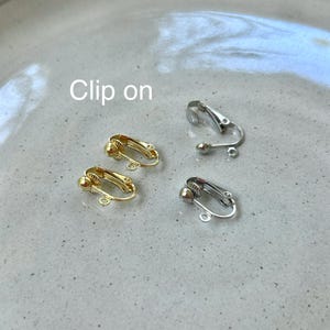 Puede incluir: Dos fornituras de pendientes de clip en tono dorado y dos en tono plateado con un peque&ntilde;o lazo circular en la base para sujetar dijes. El texto "Clip on" es visible en la parte superior izquierda de la imagen.