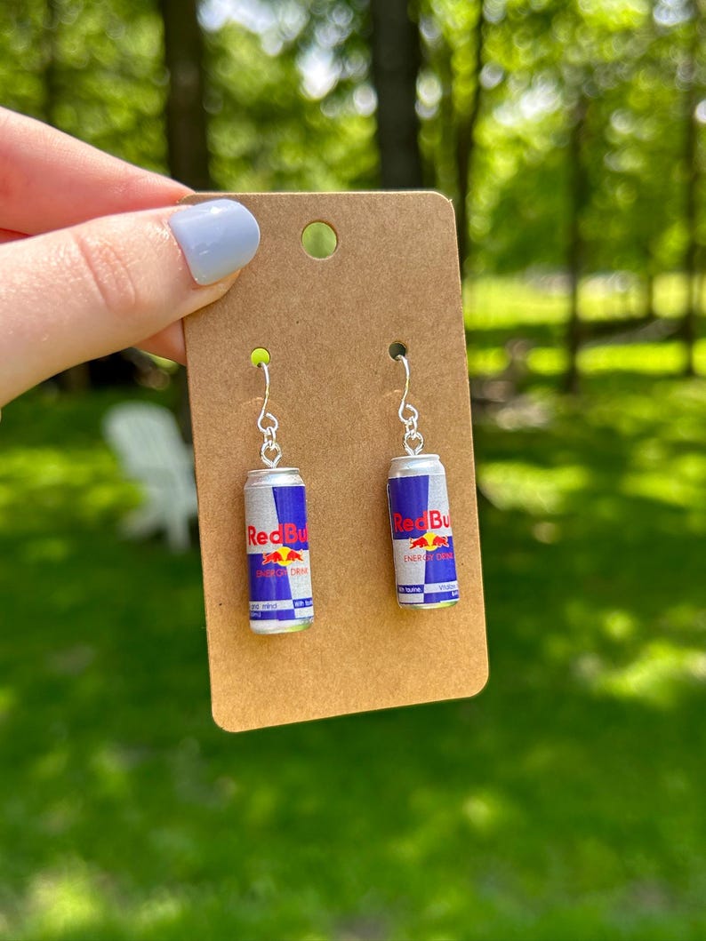 Puede incluir: Un par de pendientes colgantes con dijes en miniatura de latas de bebida energ&eacute;tica Red Bull en colores plata, azul y rojo. Los dijes est&aacute;n sujetos a ganchos de metal plateado y se presentan sobre una tarjeta de cart&oacute;n marr&oacute;n para joyer&iacute;a.