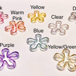 May include: A collection of ten translucent flower-shaped beads in various colors, each labeled with text: Pink, Warm Pink, Light Purple, Purple, Yellow, Blue, Clear, Dark, Green, and Yellow/Green. These small, five-petaled charms have a smooth finish.