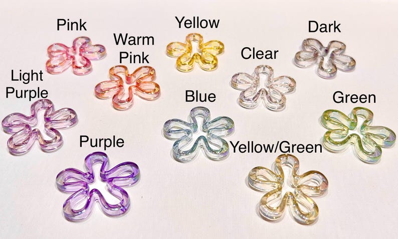 May include: A collection of ten translucent flower-shaped beads in various colors, each labeled with text: Pink, Warm Pink, Light Purple, Purple, Yellow, Blue, Clear, Dark, Green, and Yellow/Green. These small, five-petaled charms have a smooth finish.