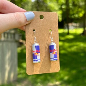 Puede incluir: Un par de pendientes colgantes con latas en miniatura de color plateado con etiquetas azules, plateadas y rojas que dicen Red Bull Energy Drink. Los pendientes est&aacute;n sujetos a una tarjeta de presentaci&oacute;n de cart&oacute;n marr&oacute;n rectangular.