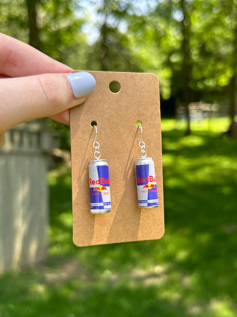 Puede incluir: Un par de pendientes colgantes con latas en miniatura de color plateado con etiquetas azules, plateadas y rojas que dicen Red Bull Energy Drink. Los pendientes est&aacute;n sujetos a una tarjeta de presentaci&oacute;n de cart&oacute;n marr&oacute;n rectangular.