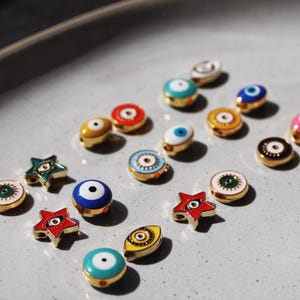 May include: A collection of colorful, gold-toned enamel charms, including round and star shapes, featuring eye designs. The charms are in various colors, such as blue, red, yellow, and green, and are arranged on a light gray surface.