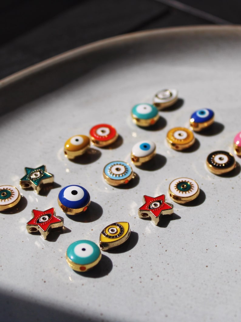 May include: A collection of colorful, gold-toned enamel charms, including round and star shapes, featuring eye designs. The charms are in various colors, such as blue, red, yellow, and green, and are arranged on a light gray surface.