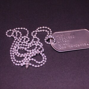 May include: A silver dog tag necklace with a silver chain. The dog tag has the text "S/N 12201990  LITCH  K.N.  DOB 10-08-1947" engraved on it.