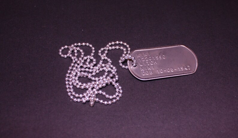 May include: A silver dog tag necklace with a silver chain. The dog tag has the text "S/N 12201990  LITCH  K.N.  DOB 10-08-1947" engraved on it.