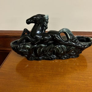 Op de afbeelding: Een zwarte keramische plantenbak met een paardenfiguur. Het paard is afgebeeld in liggende positie met het hoofd omhoog, en de plantenbak heeft een trogvorm. De glanzende afwerking en het gedetailleerde ontwerp suggereren een vintage of antieke stijl.