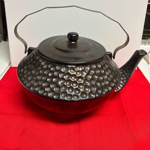 Può includere: Una teiera grigio scuro con una trama martellata, un coperchio e un manico curvo. La teiera ha un beccuccio e poggia su una superficie rossa. La teiera è in metallo.