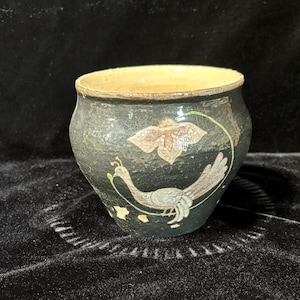 May include: A small, dark green ceramic pot with a light brown rim. The pot is decorated with a stylized peacock design in white and green, and a flower in white and brown.