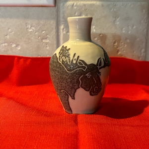 May include: A small, ceramic vase with a cream-colored body and a dark gray moose design. The vase has a narrow neck and a rounded base. The moose is depicted in a stylized, graphic manner. The vase is sitting on a red surface.