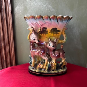May include: A vintage ceramic vase with a scalloped rim and two deer figurines. The vase is decorated with a colorful landscape scene of mountains, trees, and a sunset.