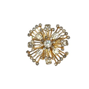 May include: A gold tone brooch with a large clear rhinestone in the center and smaller rhinestones surrounding it. The brooch is shaped like a flower with many points.