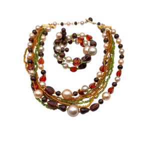 Könnte beinhalten: Ein mehrreihiges Halsketten- und Armbandset aus braunen, grünen, orangen und cremefarbenen Perlen. Die Halskette ist mit Perlen unterschiedlicher Größe geschichtet, das Armband ist ein einzelner Perlenstrang.