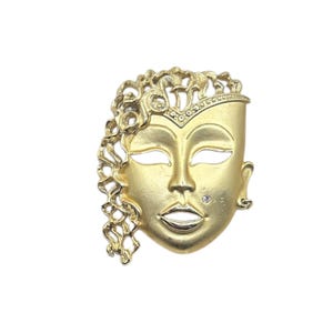 May include: A gold-toned mask-shaped brooch with a face design. The mask features open eye and mouth cutouts, a small gem on the cheek, and a decorative curly hair design on the left side. The brooch has a pin closure on the back.