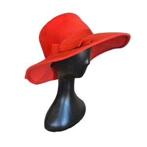 Puede incluir: Un sombrero rojo vibrante con ala ancha y lazo decorativo. El sombrero se exhibe sobre una cabeza de maniquí negra sobre un fondo blanco. El ala del sombrero mide aproximadamente 10 cm de ancho.
