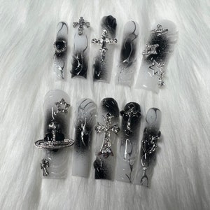 May include: A set of long, square-shaped artificial nails. Each nail features a white base with a black gradient design. Some nails are adorned with silver charms, including crosses, stars, and a heart.