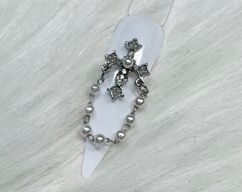 May include: A silver cross charm with a pearl and a chain of small pearls. The charm is attached to a white nail.