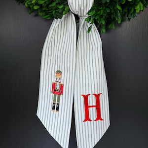 Puede incluir: Una corona de boj verde con una cinta a rayas blancas y verdes. La cinta presenta una letra "H" roja bordada y un soldado cascanueces. La corona está colgada en una puerta negra.