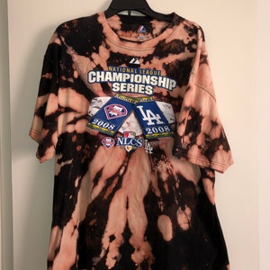Puede incluir: Una camiseta de color negro y blanco con efecto tie-dye con el texto "National League Championship Series" y los logotipos de los Philadelphia Phillies y los Los Angeles Dodgers. Los logotipos son de la temporada 2008.