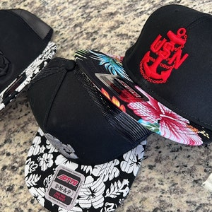 May include: Three black baseball caps with a floral print on the underside of the brim. One cap has a red embroidered anchor and the text "USN" on the front. Another cap has a black embossed anchor and the text "USN" on the front. The third cap has a white and black floral print on the underside of the brim.