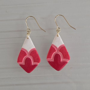 May include: A pair of handmade, drop earrings. Each earring features a white top section and a red, diamond-shaped bottom with a lighter red, curved design. The earrings have gold-colored ear wires.