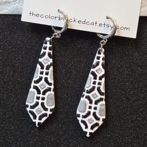 May include: A pair of dangle earrings with a geometric design. The earrings are white with black and gray accents. The earrings are hanging from silver hoops. The text "thecolorblackedcat.etsy.com" is visible on a white card.