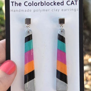 May include: Colour-blocked polymer clay earrings with silver tops and bottoms, and a colour block design of black, orange, pink, teal, and silver. The earrings are displayed on a white card with the text "The Colorblocked CAT".