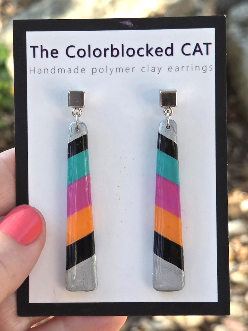 May include: Colour-blocked polymer clay earrings with silver tops and bottoms, and a colour block design of black, orange, pink, teal, and silver. The earrings are displayed on a white card with the text "The Colorblocked CAT".