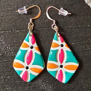 May include: A pair of teardrop-shaped dangle earrings featuring a white base with a geometric pattern in vibrant teal, bright orange, and hot pink. Each earring includes a small black dot accent and is attached to a gold-toned metal hook with a clear plastic backing.