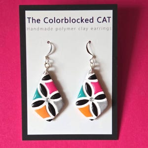 May include: Handmade polymer clay earrings in a diamond shape, featuring a colorful geometric design. The earrings have a silver hook and a flower-like pattern with black, pink, teal, orange, and gray segments. The text "The Colorblocked CAT" is visible.