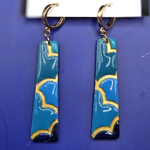 May include: A pair of gold-tone hoop earrings with rectangular, teal and blue pendants. The pendants feature a gold-colored, abstract design. The earrings are displayed against a blue background.