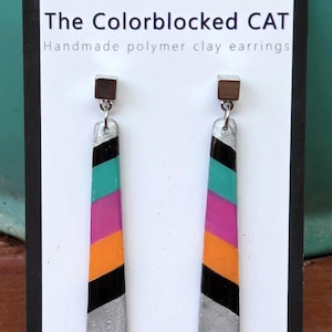 May include: Handmade polymer clay earrings with a colour-blocked design. The earrings are rectangular with silver bottoms and tops. The colour blocks include black, teal, pink, orange, and black. The text on the card reads "The Colorblocked CAT".