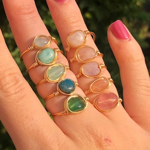 Op de afbeelding: Een set van acht gouden draadgewikkelde ringen met verschillende gekleurde edelstenen. De ringen zijn gestapeld op een hand, met de edelstenen naar de kijker gericht. De edelstenen zijn groen, blauw, roze en helder.