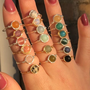May include: A set of 12 gold and silver wire wrapped rings with different coloured gemstone beads. The rings are stacked on a hand.