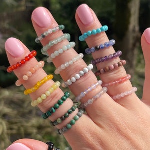 Op de afbeelding: Een hand met een verscheidenheid aan gekleurde kralenringen. De ringen zijn gemaakt van verschillende soorten stenen, waaronder turquoise, amethist en malachiet.