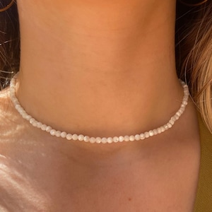 Könnte beinhalten: Eine zarte, weiße Perlenkette. Die Halskette besteht aus kleinen, runden Perlen, die einen dezenten, eleganten Choker-Stil bilden. Die Perlen scheinen eine helle, perlmuttartige weiße Farbe zu haben und reflektieren das Licht.