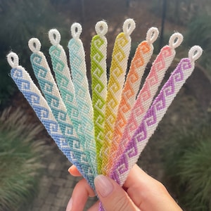 Pode incluir: Um conjunto de oito pulseiras de amizade em um padrão de cores do arco-íris. As pulseiras são feitas com fio branco e apresentam um design geométrico.