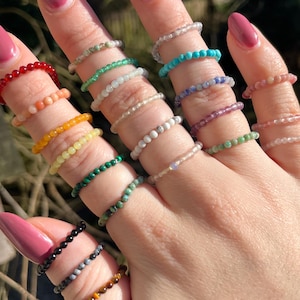 Op de afbeelding: Een hand met een verscheidenheid aan edelsteenringen. De ringen zijn gemaakt van kleine kralen en zijn in een regenboog van kleuren.