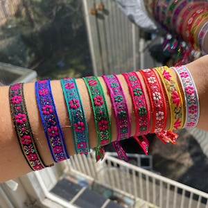 May include: A set of ten colourful woven bracelets with pink flower designs. The bracelets are stacked on a person's arm and feature a variety of colours, including black, blue, green, red, yellow, and white.