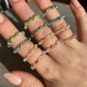Könnte beinhalten: Eine Hand zeigt mehrere Perlenringe in verschiedenen Farben, darunter Grün, Blau, Pfirsich und Rosa. Die Ringe sind auf einem Gummiband aufgefädelt und verfügen über kleine, facettierte Perlen mit goldfarbenen Akzenten. Die Ringe sind ein zartes und farbenfrohes Accessoire.