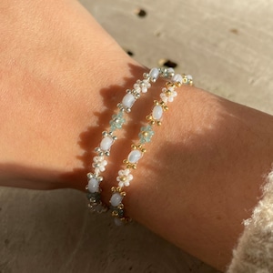 Op de afbeelding: Twee delicate kralenarmbanden met witte en lichtblauwe kralen. De armbanden zijn gemaakt met kleine, ronde kralen en hebben een bloemmotief.