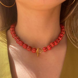 Op de afbeelding: Een rode kralenketting met een goudkleurige sluiting. De ketting heeft grote, ronde kralen in een levendige rode kleur. De sluiting is een eenvoudig staaf- en lusontwerp. De ketting wordt om de nek gedragen.