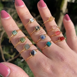 Puede incluir: Un maniquí de mano con una variedad de anillos de oro con piedras preciosas de diferentes colores. Los anillos están hechos con alambre de oro fino y presentan piedras preciosas de forma cuadrada.