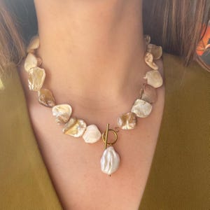 Op de afbeelding: Een opvallende ketting bestaande uit onregelmatig gevormde, iriserende schelpstukken in crème-, bruin- en bruintinten. Het heeft een goudkleurige sluiting en een grote, peervormige parelhangert.