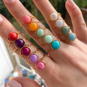 Op de afbeelding: Een verzameling handgemaakte ringen met gouden draadzettingen. Elke ring heeft een kleurrijke, ronde edelsteen in tinten rood, roze, oranje, groen, paars en blauw. De ringen worden op een hand getoond, waarbij de verscheidenheid aan kleuren en ontwerpen wordt getoond.