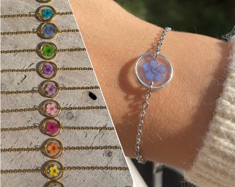 Könnte beinhalten: Ein silbernes Kettenarmband mit einem zarten Blumenanhänger. Die Blume ist gepresst und in einem klaren Harz-Kreis konserviert. Das Armband wird am Handgelenk einer Person gezeigt.