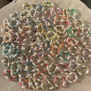 Puede incluir: Una colecci&oacute;n de anillos de cuentas coloridas con cuentas blancas y detalles dorados. Los anillos est&aacute;n dispuestos en un patr&oacute;n circular sobre una superficie marr&oacute;n claro.