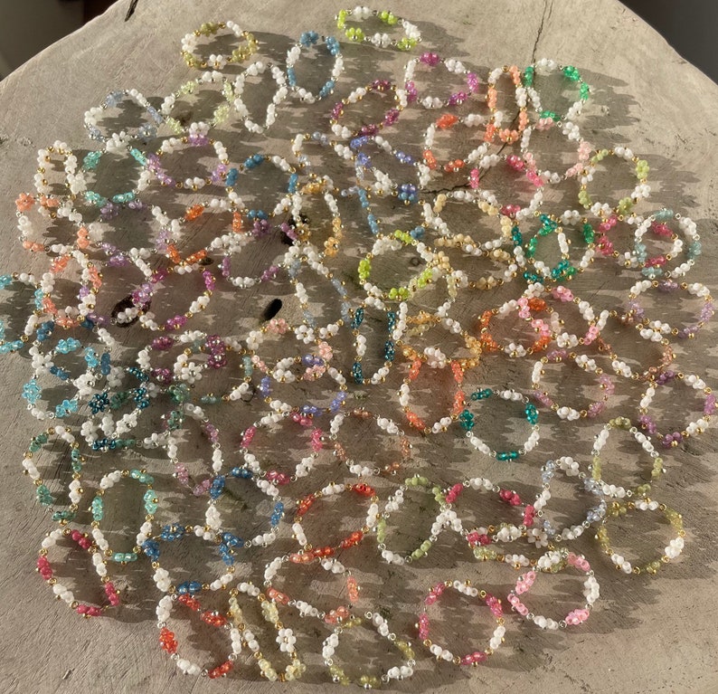Puede incluir: Una colecci&oacute;n de anillos de cuentas coloridas con cuentas blancas y detalles dorados. Los anillos est&aacute;n dispuestos en un patr&oacute;n circular sobre una superficie marr&oacute;n claro.